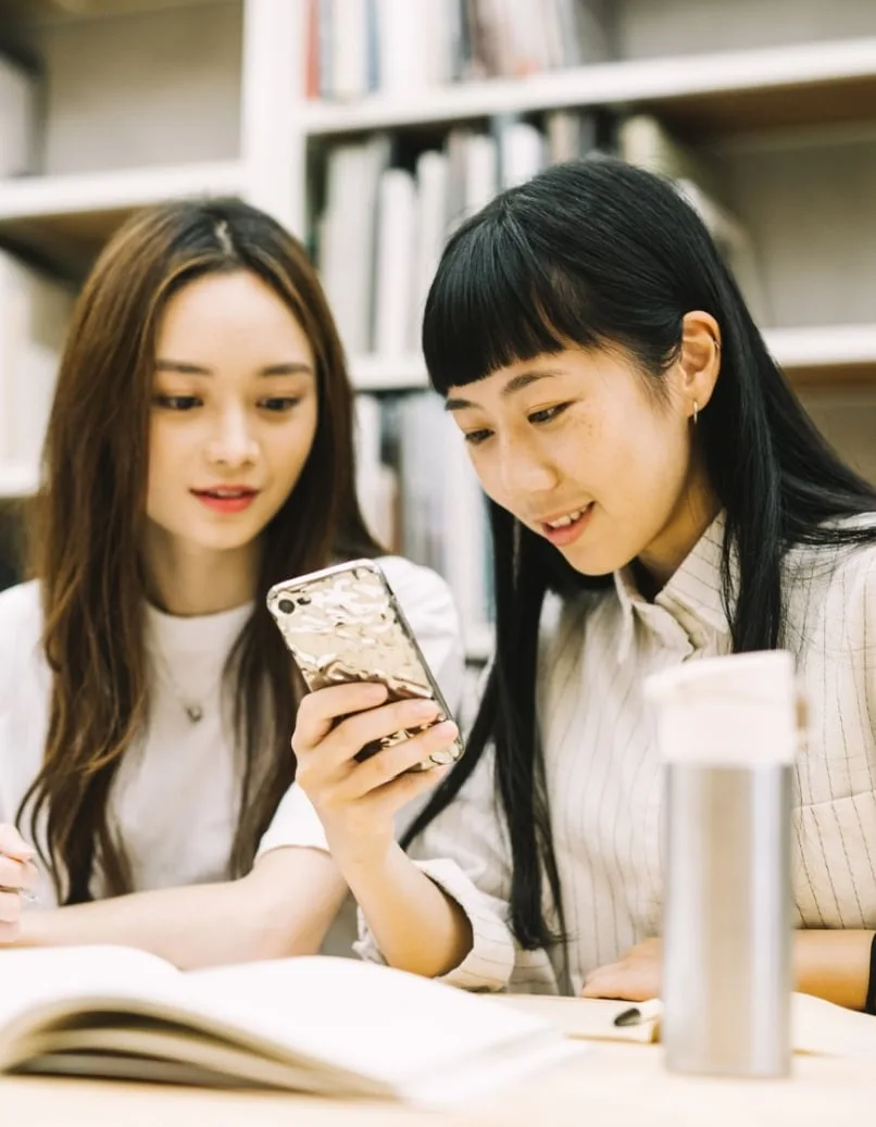 写真：図書館で一緒にスマートフォンを見る2人のアジア人女性学生