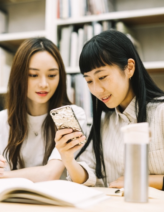 写真:2人の女性がテーブルの前でスマホを見ながら笑顔で会話している。